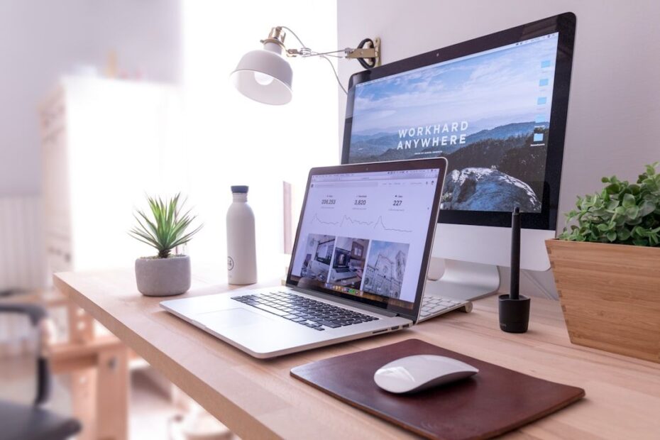 A modern digital workplace setup with a laptop, desktop monitor, and mouse on a wooden desk.